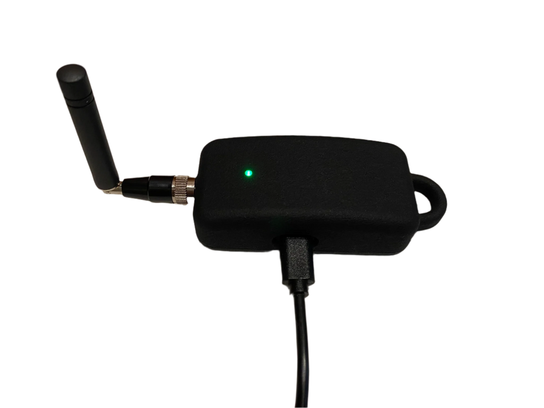 The SkyShepherd mobile gateway is plugged into a charging cord. The attachable antenna is bent at the integrated elbow at a 90 degree angle, perpendicular to the ground. The gateway's LED light is illuminated and green. The integrated carry loop can be seen on the side of the gateway opposite the antenna. The gateway sits in it's normal plugged-in orientation, resting horizontally on an unseen surface.